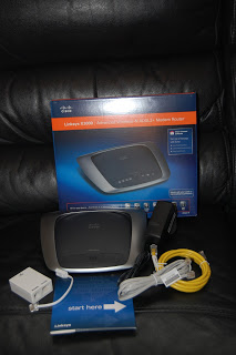 A Linksys E1200 wireless router, featured in this Router Review, comes with its box, power adaptor, yellow Ethernet cable, white adaptor, and instruction manual neatly arranged on a black surface.
