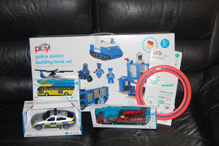 Part of the Wilkinson Toy Range, this set features a police station brick set, toy police car, helicopter, train, and a red circular track—all displayed on a black leather sofa.