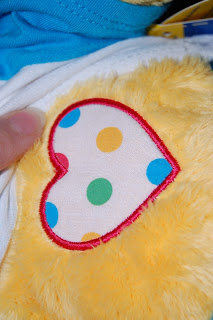 A close-up of a yellow plush toy from Build-A-Bear Workshop with a white heart patch decorated with colourful polka dots and outlined in red. A finger is gently touching the patch, symbolising support for children in need.