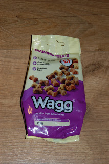 A bag of Wagg Training Treats for dogs on a wooden surface. The purple and yellow packaging features images of the Dog Treats along with text highlighting the product’s health benefits.