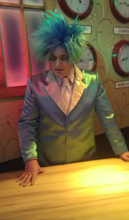 A person with bright blue spiky hair and a shiny blue suit stands behind a wooden counter in Thomas Land, looking down. Several large clocks are mounted on the wall behind them, adding to the Magical Christmas atmosphere.