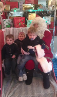 Three young children sit with Father Christmas on a red chair at Drayton Manor, surrounded by wrapped Christmas presents. One child has a dummy, and all are dressed warmly for the Magical Christmas experience.