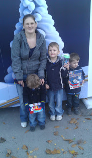 A woman stands behind three young boys in jackets at Drayton Manor. Two boys hold toys whilst leaves are scattered on the ground, and a large, white, tree-like decoration hints at a Magical Christmas in Thomas Land.