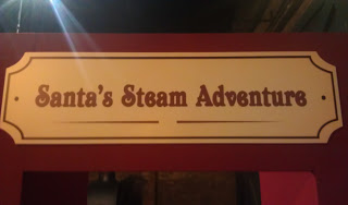 A cream-coloured sign with a decorative border reads "Santa's Christmas Steam Adventure" in brown text. The sign is mounted on a red background at the National Railway Museum, York.