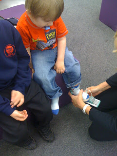 A young child in a red shirt sits as an adult helps put a plaster on his ankle, possibly after removing his Clarks Shoes. Another child in a navy sweatshirt sits beside him indoors on a carpeted floor.