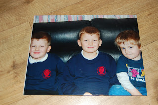 A Snapfish printed photo shows three young children on a black sofa—two in matching navy blue school uniforms, one in a cartoon t-shirt. The photo rests on a light wooden floor.