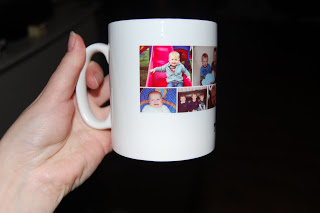 A hand holds a white mug from Snapfish, decorated with four small photos of a baby and young child in various poses and settings. The background is dark and out of focus.