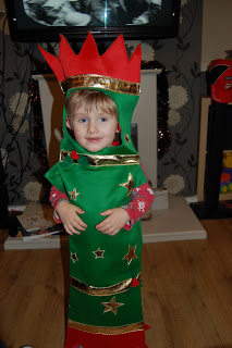 A young child smiles whilst wearing a green and red Christmas tree costume from Fancy Dress Outfitters, decorated with gold stars and trim, standing indoors on a wooden floor.