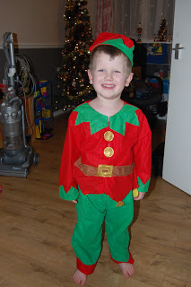 A young child smiles whilst wearing a festive elf costume from Fancy Dress Outfitters, standing barefoot on a wooden floor with a decorated Christmas tree and vacuum cleaner in the background.