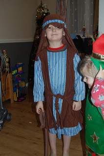 A young child wearing a blue striped dressing gown with a brown sash and head covering stands indoors, dressed as a shepherd—an ideal look from Fancy Dress Outfitters. Another child in a green starry costume and red hat is beside them, partially out of frame.