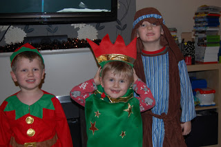 Three young children in festive costumes stand indoors, smiling after a fun Christmas Crafts challenge. One is dressed as an elf, another wears a green costume with a red crown, and the third sports a striped robe with a brown head covering.