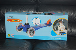A blue box on a black leather sofa contains a Mini Micro Scooter. The box features an image of the blue and orange scooter and a photo of a smiling child wearing a helmet.