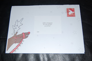 A white envelope featuring a cartoon reindeer on the left side, a red postage stamp in the upper right corner, and an address label in the centre—perfect for sending magical Letters from Father Christmas. The background is a dark surface.
