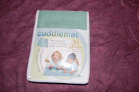 A pack of a green Cuddlemat by Cuddledry, shown on a purple surface. The label displays two happy children lying on the mat and text that reads “cuddlemat” with additional product information.