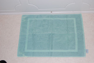 A light blue rectangular Cuddledry bath mat with a simple border design is placed on a light-coloured carpeted floor in front of a white wall.