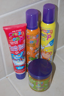 Four colourful bottles of Crazy Soap bath products for children, including blue bathtime body paint, two foam sprays, and a round jar, all with purple lids, placed on a tiled bathroom surface.