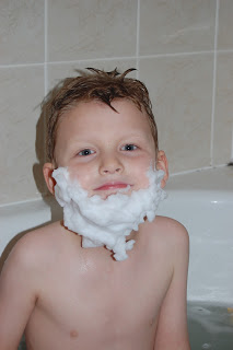 A young boy sitting in a bath with wet hair and a thick layer of white soap bubbles shaped like a beard on his face, smiling slightly at the camera.