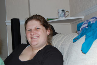 A determined woman with light brown hair tied back is smiling whilst sitting on a light-coloured sofa. Behind her, a mug and some folded clothes are visible on a white shelf, reflecting her focus on losing weight.