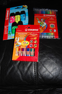 Three packs of Stabilo writing products—coloured pencils, highlighters with animal faces, and a set of coloured pens—lay on a black leather surface.