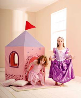 Two young girls dressed as princesses play in a room; one stands in a purple gown and tiara, while the other exits a Fairy Princess Play Tent with a red flag. Light streams through a window, and two pillows are on the floor.