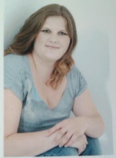 A young woman with long, wavy brown hair, wearing a light blue short-sleeved shirt, sits with her arms resting on her knees and smiles softly at the camera, radiating confidence from her recent weight loss journey against a plain light background.