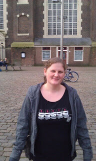 A woman with light brown hair stands smiling on a cobbled square in front of a large brick building with tall windows, radiating confidence after her weight loss journey. A blue bicycle is parked nearby, and a bench is visible in the background.