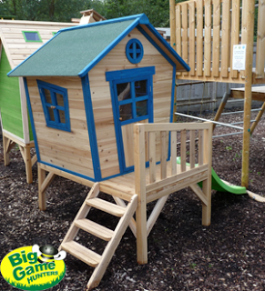 A small wooden playhouse with blue trim, a green roof, steps, and a cosy porch rests on bark mulch. With a green slide attached and "Big Game Hunters" logo visible, it’s perfect for imaginative fun or as inspiration for your next Fairy Princess Play Tent!.