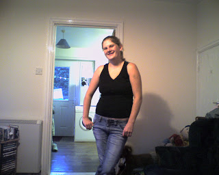 A woman in a black sleeveless top and jeans stands smiling in a living room, showing her confidence after losing weight. One arm rests on her hip, with a doorway behind her leading to a kitchen area with visible appliances.