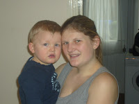 A young woman with long brown hair, determined to win at losing weight, smiles while holding a small child with blonde hair and a blue shirt indoors, near a window with white curtains.