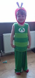 A young child stands indoors, smiling in a green and yellow striped costume with a red headpiece resembling The Very Hungry Caterpillar, celebrating World Book Day. A radiator can be seen in the background.