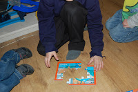 Three children sit on a wooden floor working together on a colourful Octonauts jigsaw puzzle. One child is placing a puzzle piece while the others sit nearby, watching and waiting to help complete the 4 in a box set.