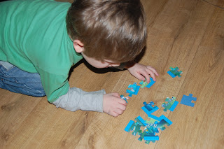 A young child in a green shirt is kneeling on a wooden floor, focusing on assembling blue Octonauts jigsaw puzzle pieces from a colourful box.