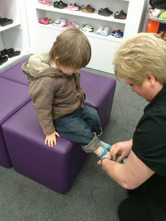 A small child sits on a purple bench in a shoe shop while an adult helps try on James’s new shoes. Shelves with various children's shoes are visible in the background.