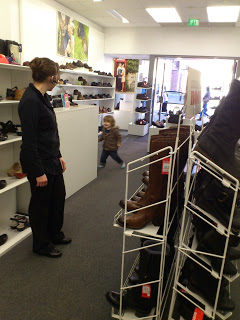 A woman stands near a display in a shoe shop, watching a small child—perhaps excited about James' New Shoes—run towards the entrance. Shelves filled with various shoes and boots line the walls and floor.