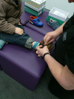 A child sits on a purple bench whilst an adult helps fit James's new shoes on the child’s foot. Other pairs of shoes and shoe boxes are visible nearby.