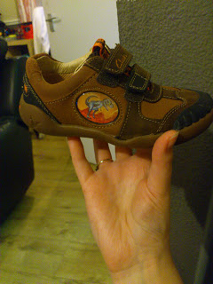 A hand holding James’s New Shoes—a small brown children’s trainer with two Velcro straps and a colourful cartoon animal patch on the side, set against a grey wall and wooden floor background.