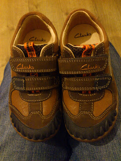A pair of brown Clarks children's shoes, dubbed James' New Shoes, with double Velcro straps and orange detailing, are placed side by side on a person's lap with blue jeans visible underneath.