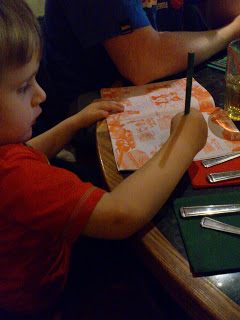 A young child in a red shirt sits at a table in Frankie & Benny's, drawing on a paper placemat with a green pencil. Cutlery and serviettes are arranged nearby.