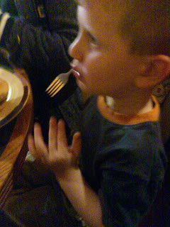 A young boy sits at a table indoors at Frankie & Benny's, holding a fork near his mouth and looking to the side, with a plate of food in front of him. The image is slightly blurry.