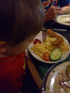 A child in a red shirt looks at a plate of food with chips, chicken nuggets, a slice of cucumber, a cherry tomato, and tomato ketchup at Frankie & Benny's. An adult is holding a fork near the plate.