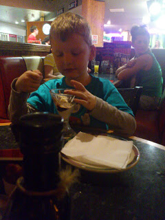 A young boy sits at a Frankie & Benny's restaurant table eating pudding from a glass dish, with serviettes on a plate in front of him. Other diners and classic Frankie & Benny's decor are visible in the background.