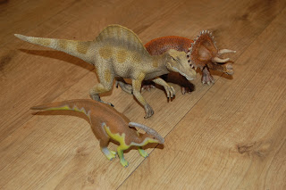 Three plastic dinosaur toys, including a Spinosaurus, Triceratops, and Parasaurolophus from Schleich Dinosaurs, are arranged on a wooden floor.