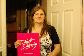 A woman with long brown hair stands indoors, smiling and holding a bright pink "Dirty Dancing" folder in front of her chest—ready to relive the Time of My Life. She wears a black and white floral top with a black cardigan.