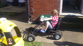 A child wearing a red and white striped shirt and colourful socks sits in a non-electric pedal go-kart on a sunny drive, near a brick building and a yellow toy car, ready to take on the Battery Challenge.