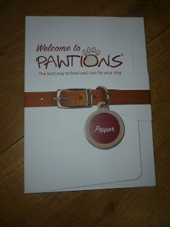 A folder with the text "Welcome to PAWTIONS: The best way to feed and care for your dog," featuring a Pawtions dog collar and a tag labelled "Pepper" on a wooden surface.