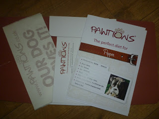 A Pawtions folder with documents, including a diet plan labelled "The perfect diet for Pepper," a dog’s photo, a red collar design, and a white slip reading "OUR DOG LOVES IT!" sits on a wooden surface.