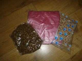 Three Pawtions plastic bags on a wooden floor: one with brown kibble, one with pink dog waste bags, and one with small dog treats in a paw-print patterned bag.