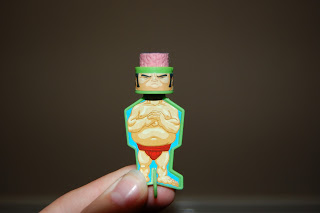 A hand holds a small Scatter Brainz toy figure of a muscular sumo wrestler with a pink textured top, angry expression, and red pants—perfect for fans of quirky boys' toys—against a plain brown background.