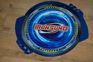 A round blue plastic toy arena with a swirling design, perfect for Monsuno Strike battles, featuring bold red and white "Monsuno" lettering at the centre and placed on a wooden floor.