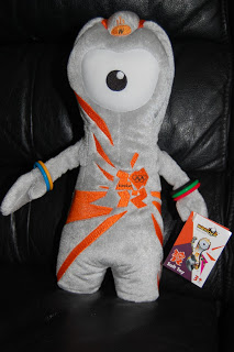 A Wenlock Soft Toy mascot from the London 2012 Olympic Games, featuring a single large eye, grey body with orange patterns, two bracelets, and holding a tag with the official Olympic logo.
